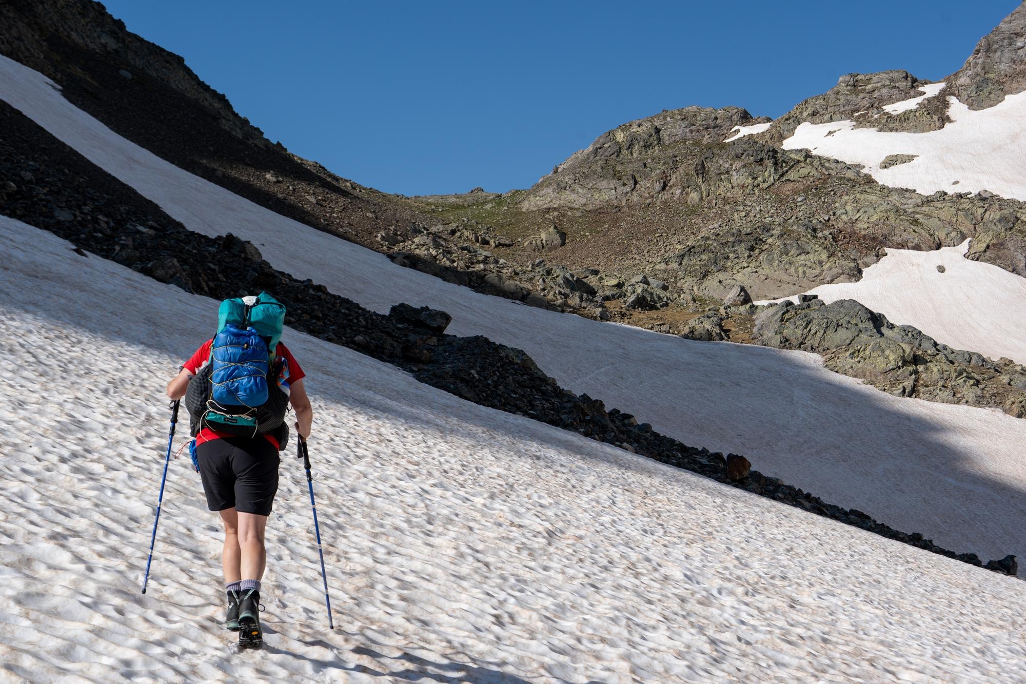 A hiker ascends a snow slope leading to a snow free mountain pass