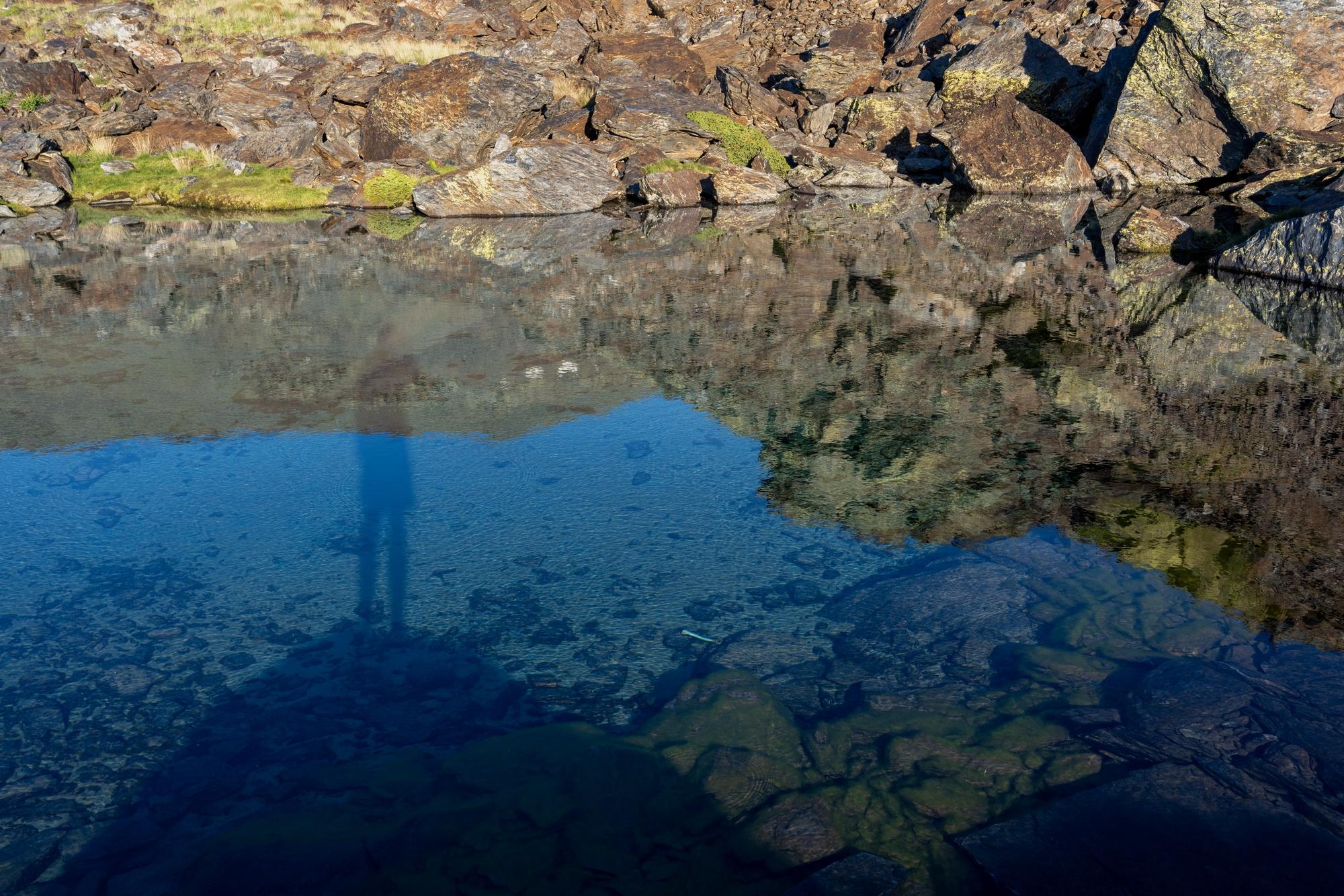 A person stood on a rock platform is reflected in the blue waters of a mountain lake
