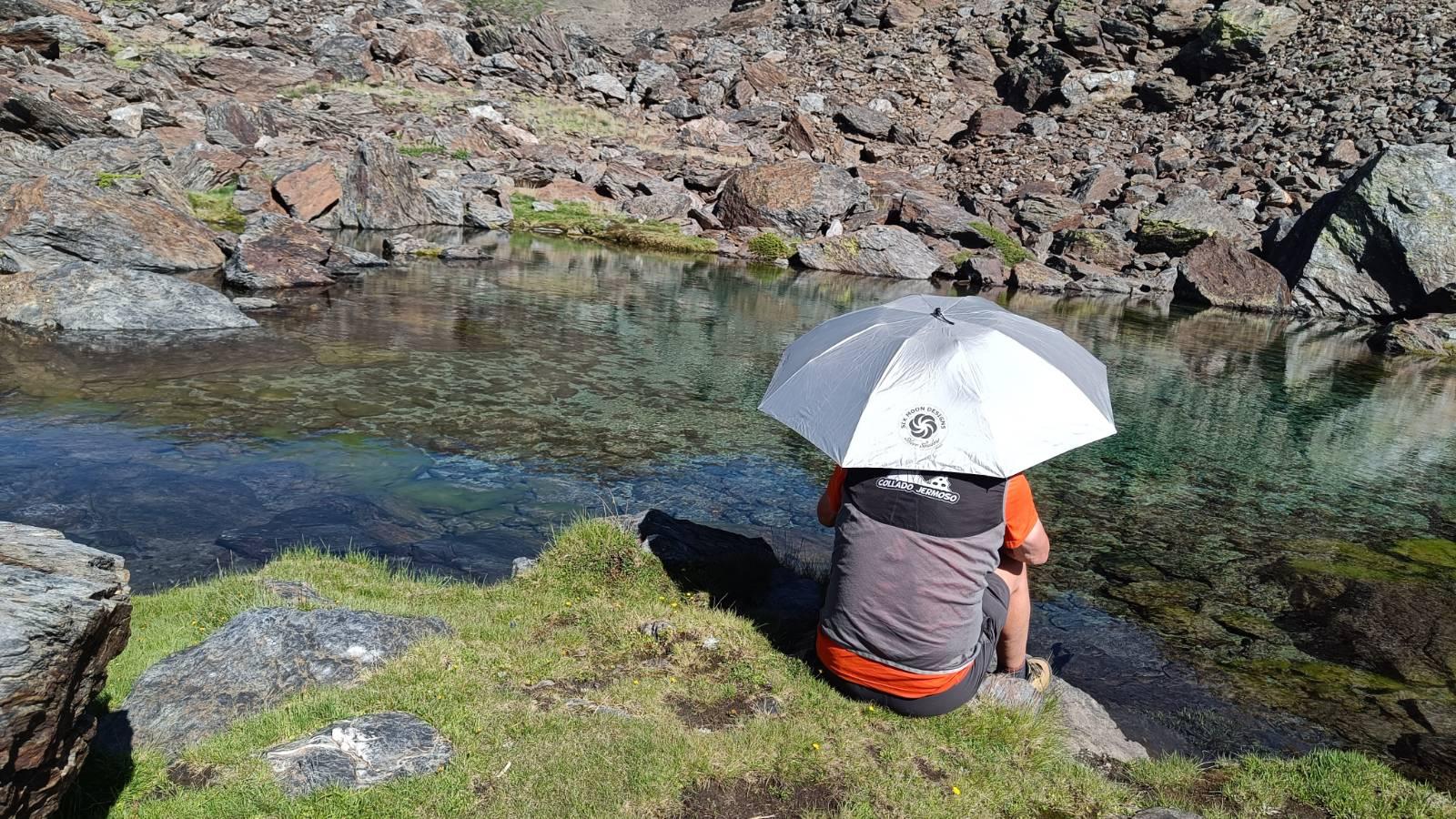 A person sits on the side of a lake holding a silver umbrella to shield himself from the suns rays