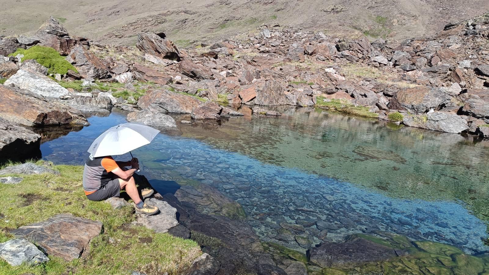 A person sits on the side of a lake holding a silver umbrella to shield himself from the suns rays