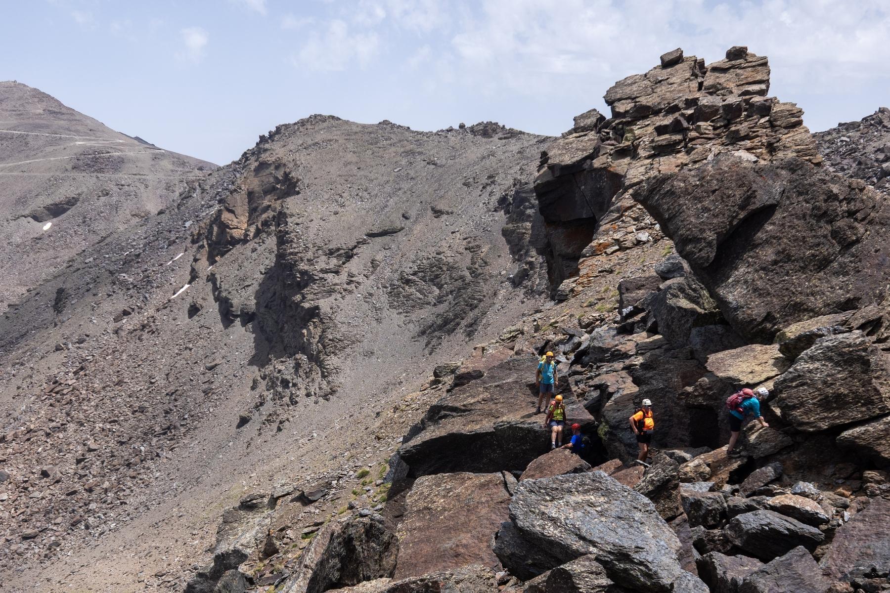 A group of people in colorful clothing and helmets pass along a boulder strewn mountain ridge