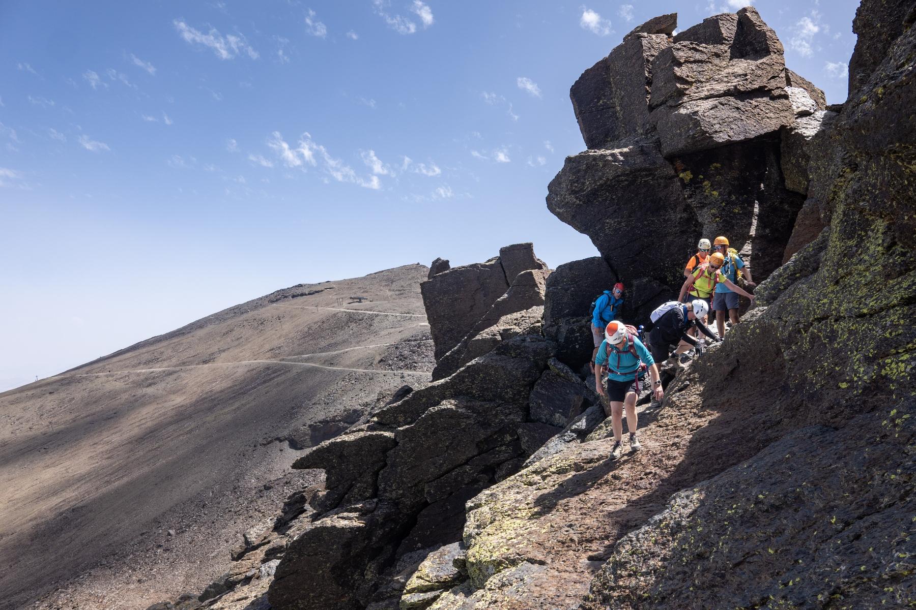 A group of people in colorful clothing and helmets pass along a narrow ledge with vertical rock above and below