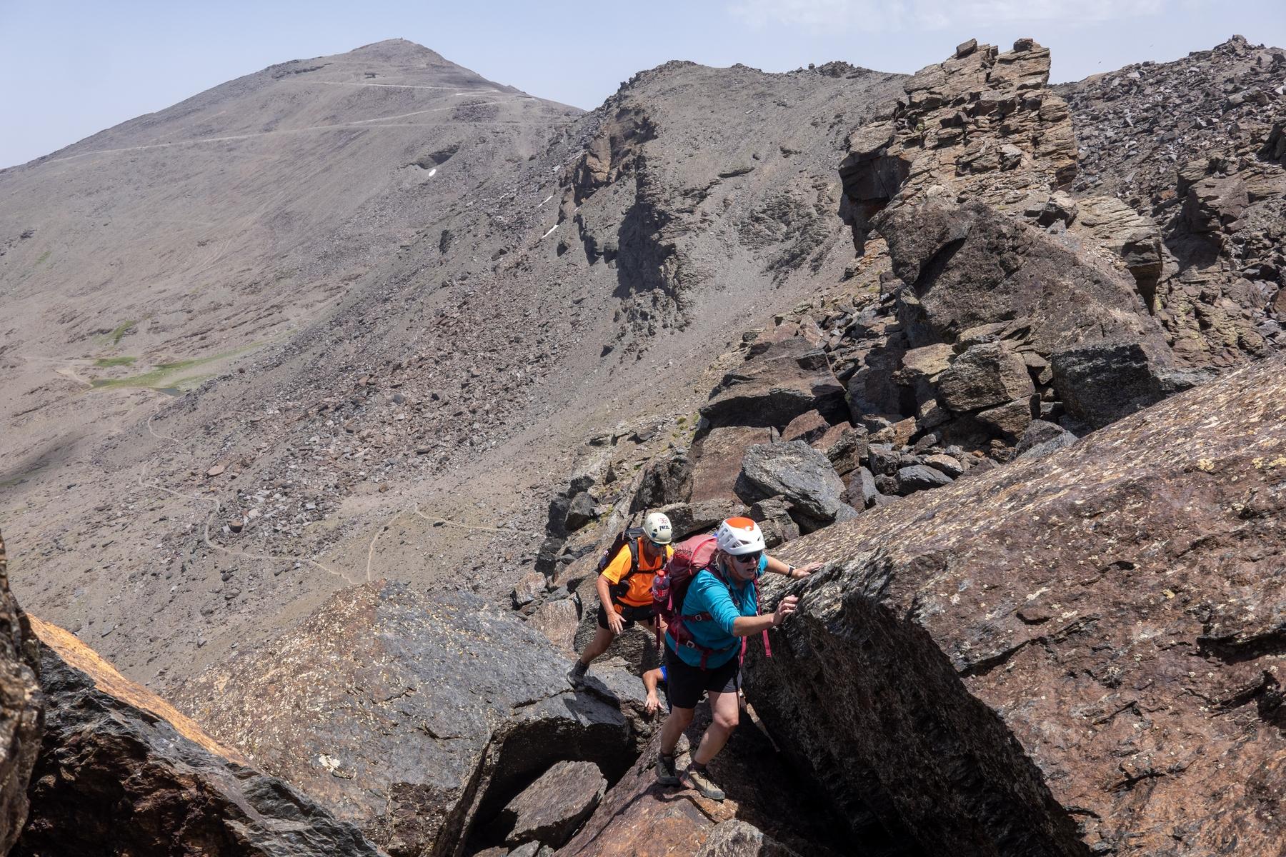 Two people ascend some rocks on a mountain ridge