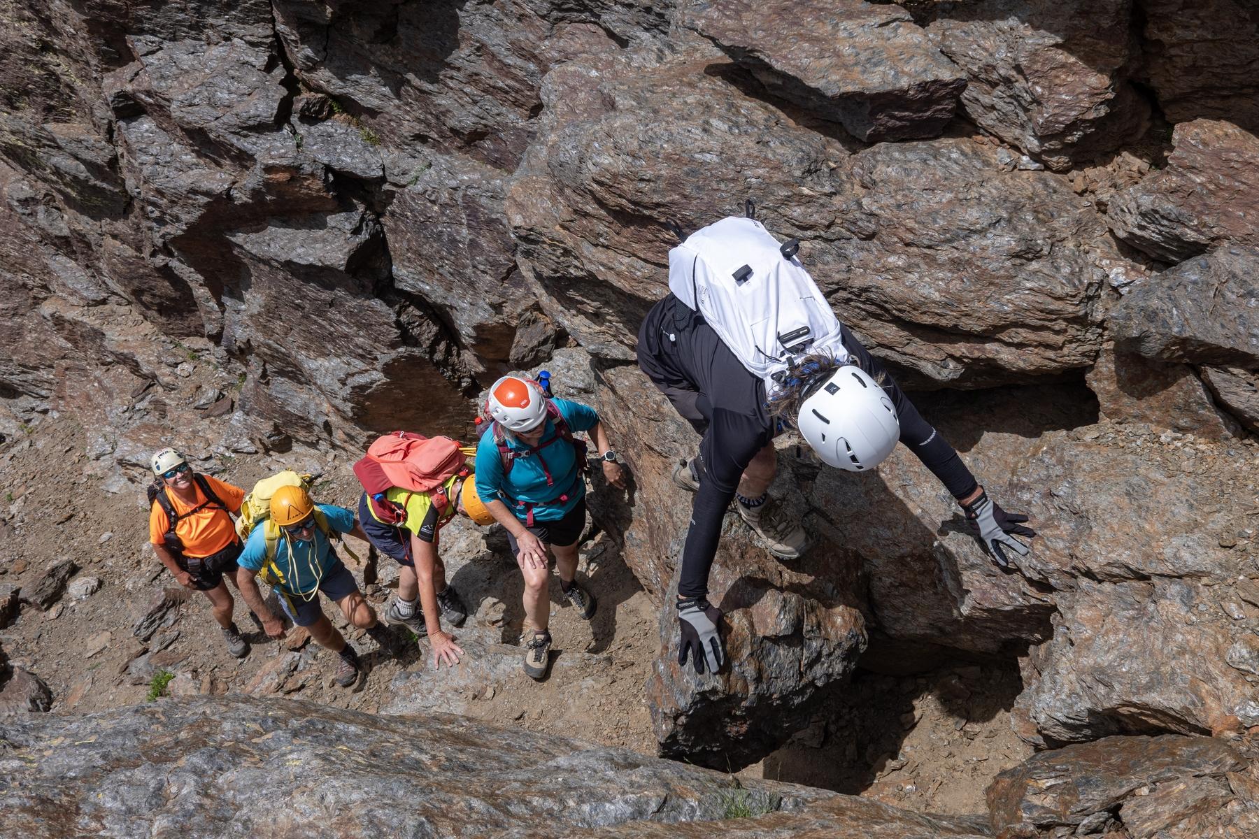 A group of helmeted hikers in colorful clothes clamber and scramble along a rocky and block strewn high level mountain ridge