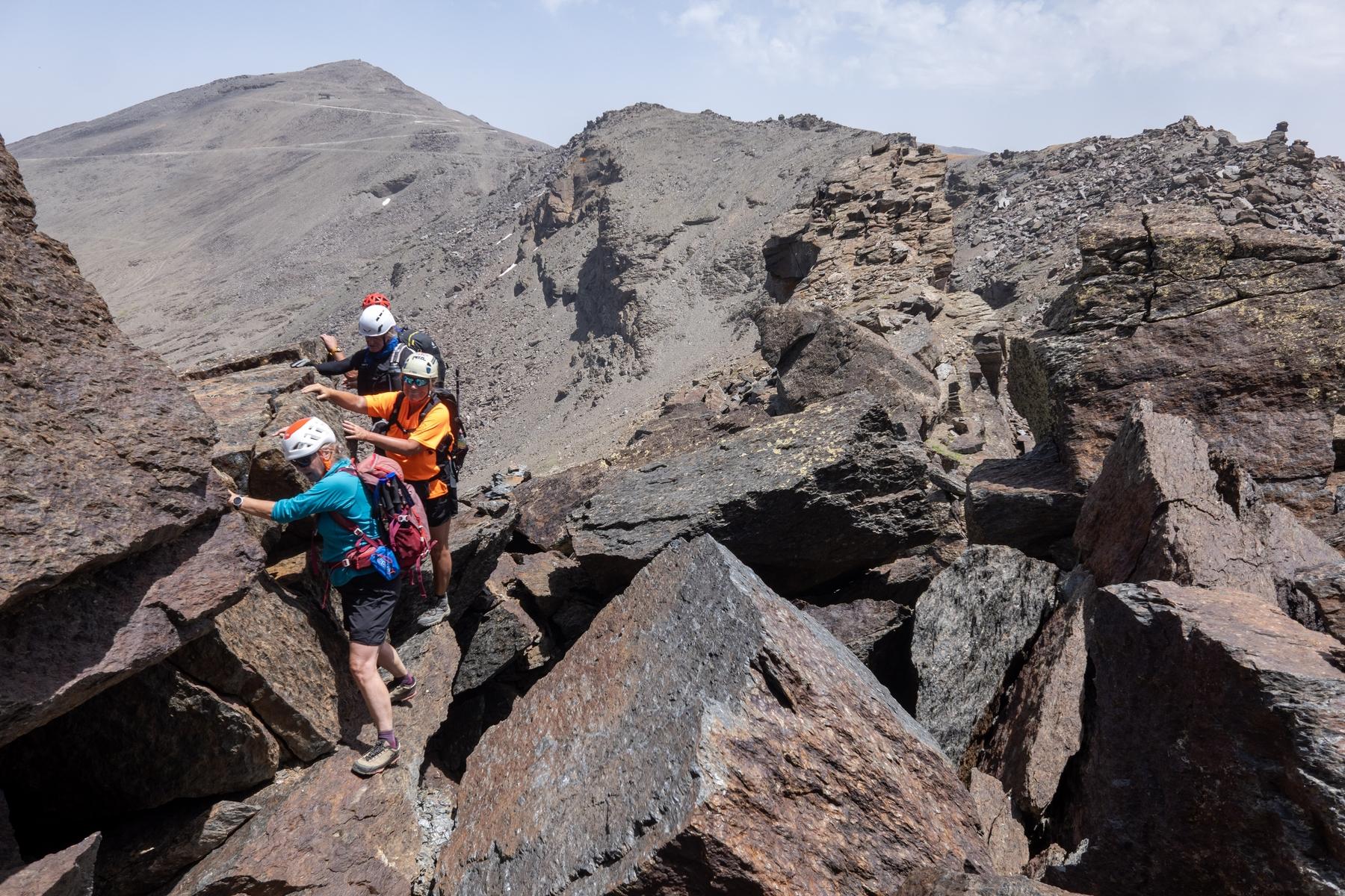 A group of helmeted hikers in colorful clothes clamber and scramble along a rocky and block strewn high level mountain ridge