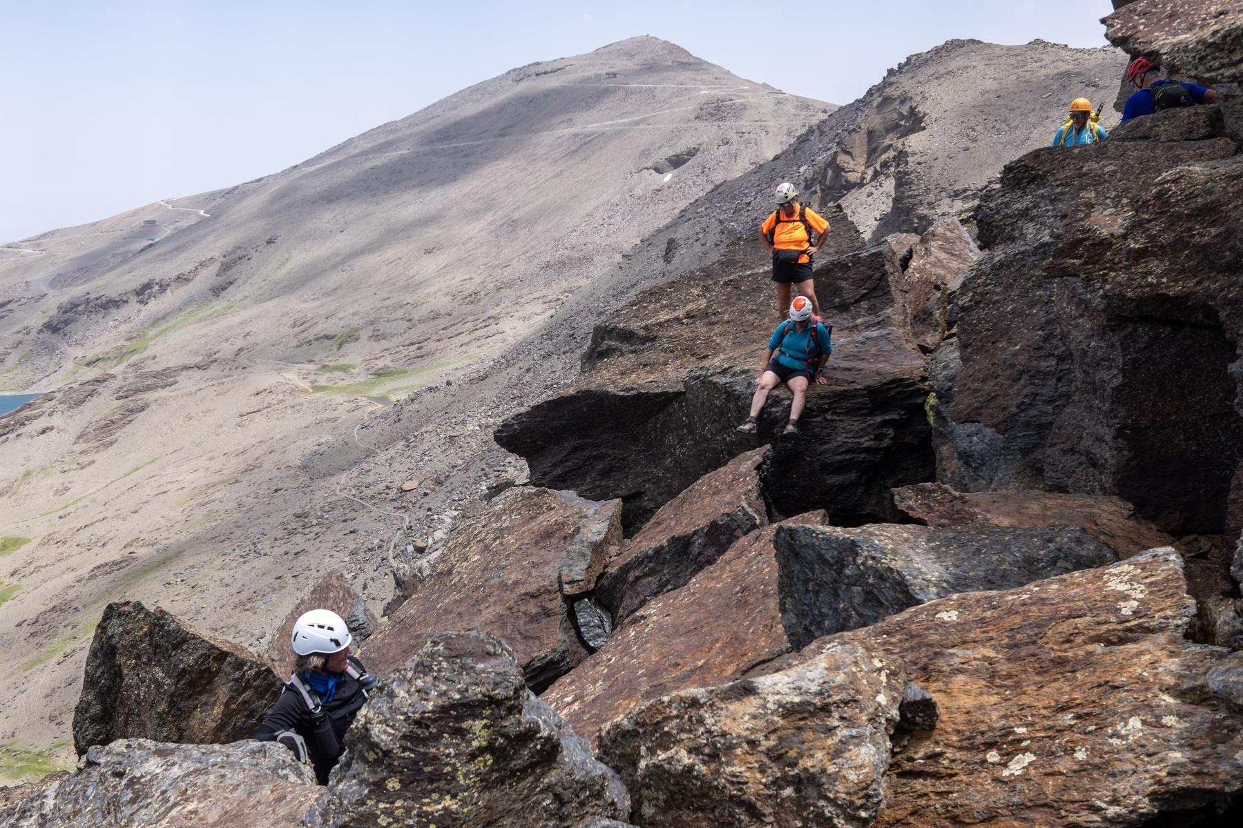 A group of helmeted hikers in colorful clothes clamber and scramble along a rocky and block strewn high level mountain ridge
