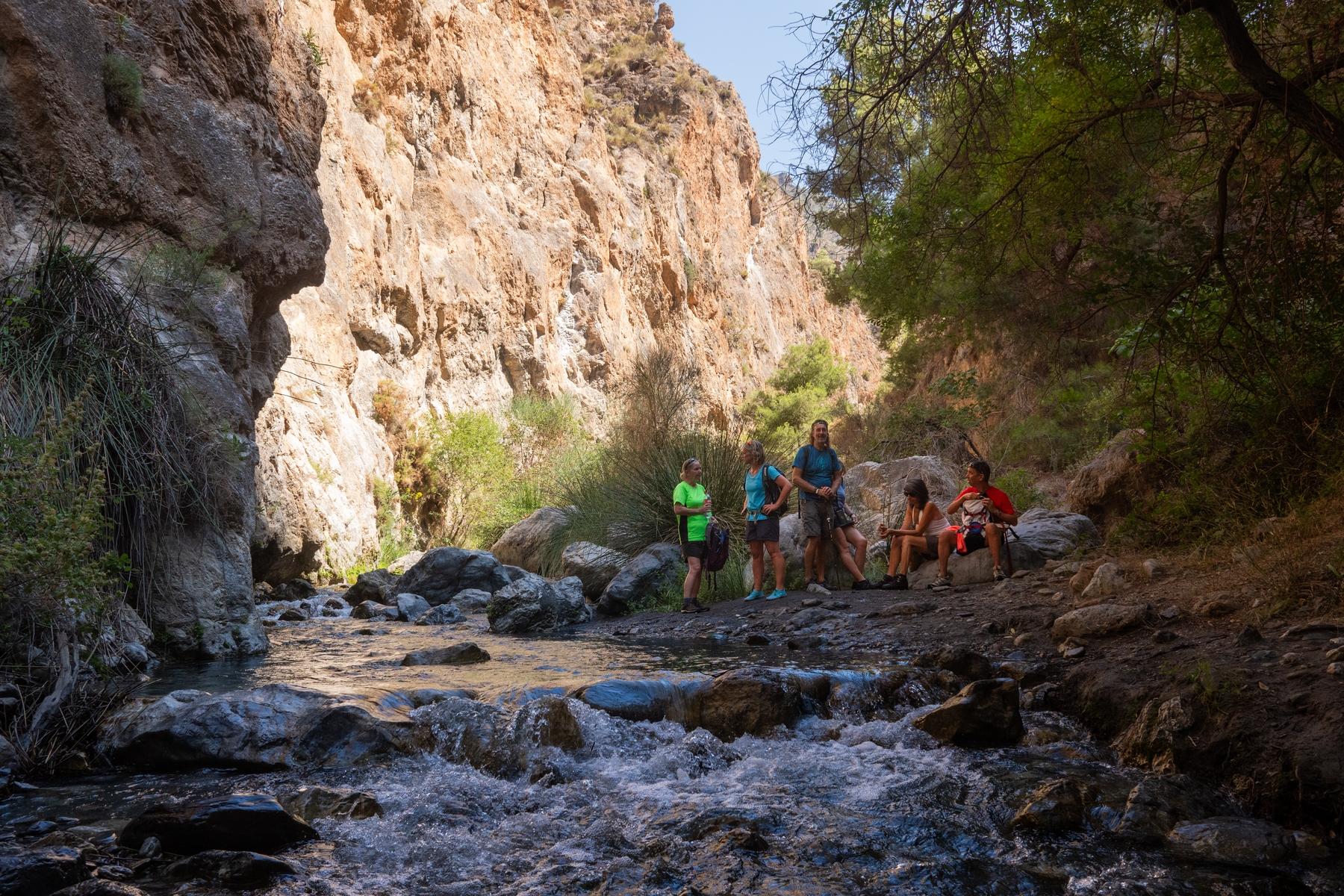 A group of hikers stands alongside a shallow river with some spectacular cliff scenery above and to the left