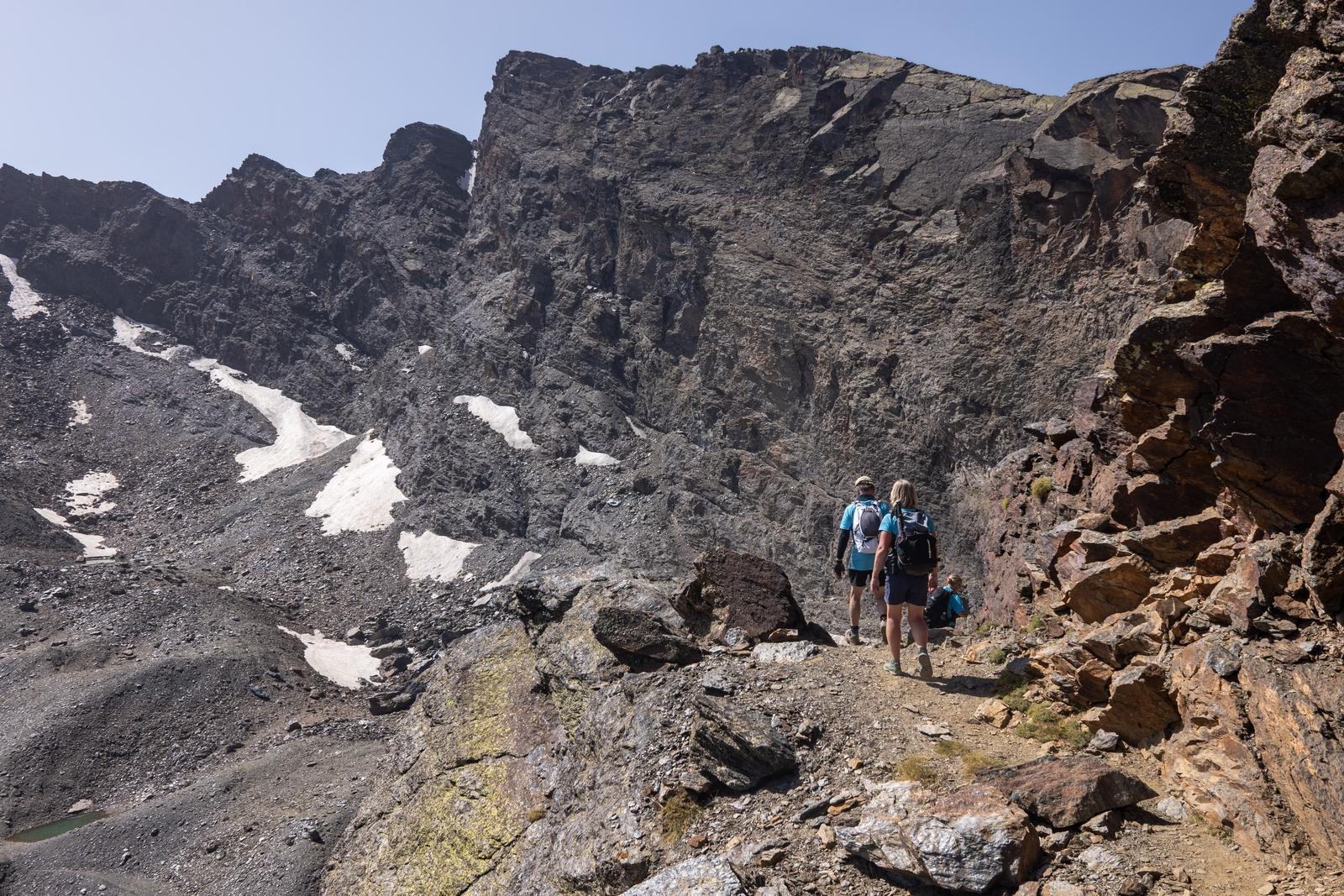 Two hikers on a narrow path are surrounded by rock walls. Ahead lies the peak of Veleta with some late season snow still around