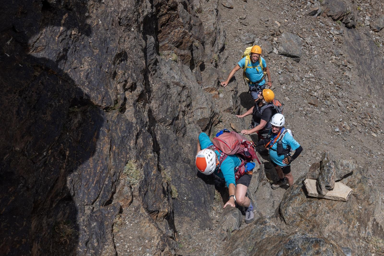 4 scramblers in brightly colored gear and helmets start ascending a short rock wall