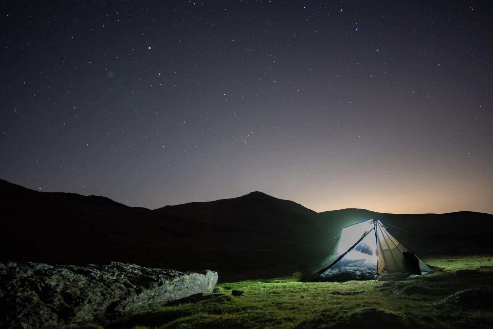 The last light of the sunset is fading and the stars appear over to the west. A tent is illuminated by headtorch. The brightness of the torch illuminates the green grasses next to the torch
