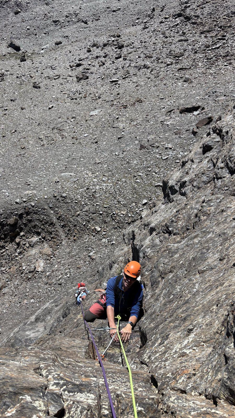 The author in a blue top and orange helmet is rock climbing up a near vertical crack system. Below can be seen another climber