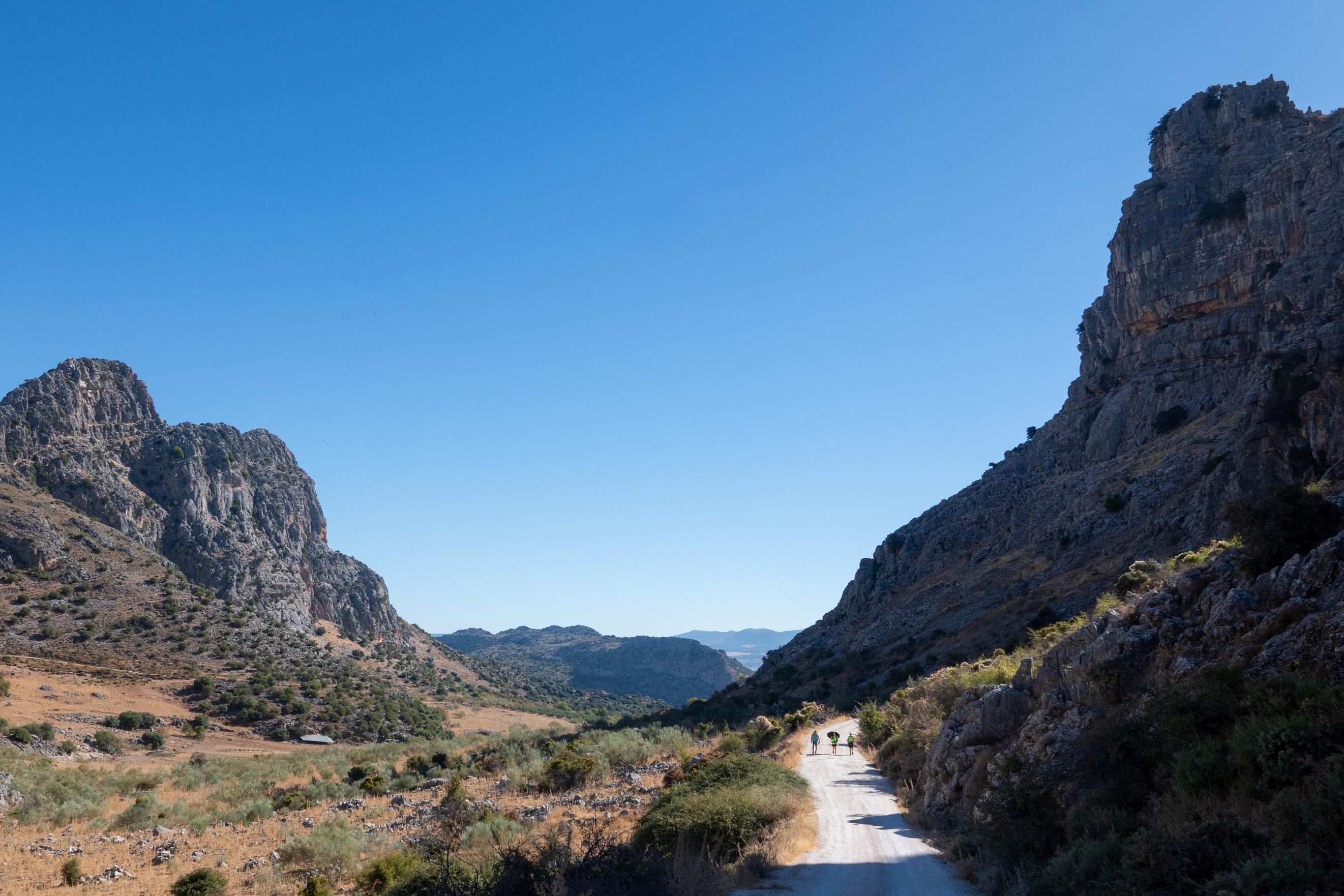 3 people pass along a wide trail with shadowed mountainside to the right