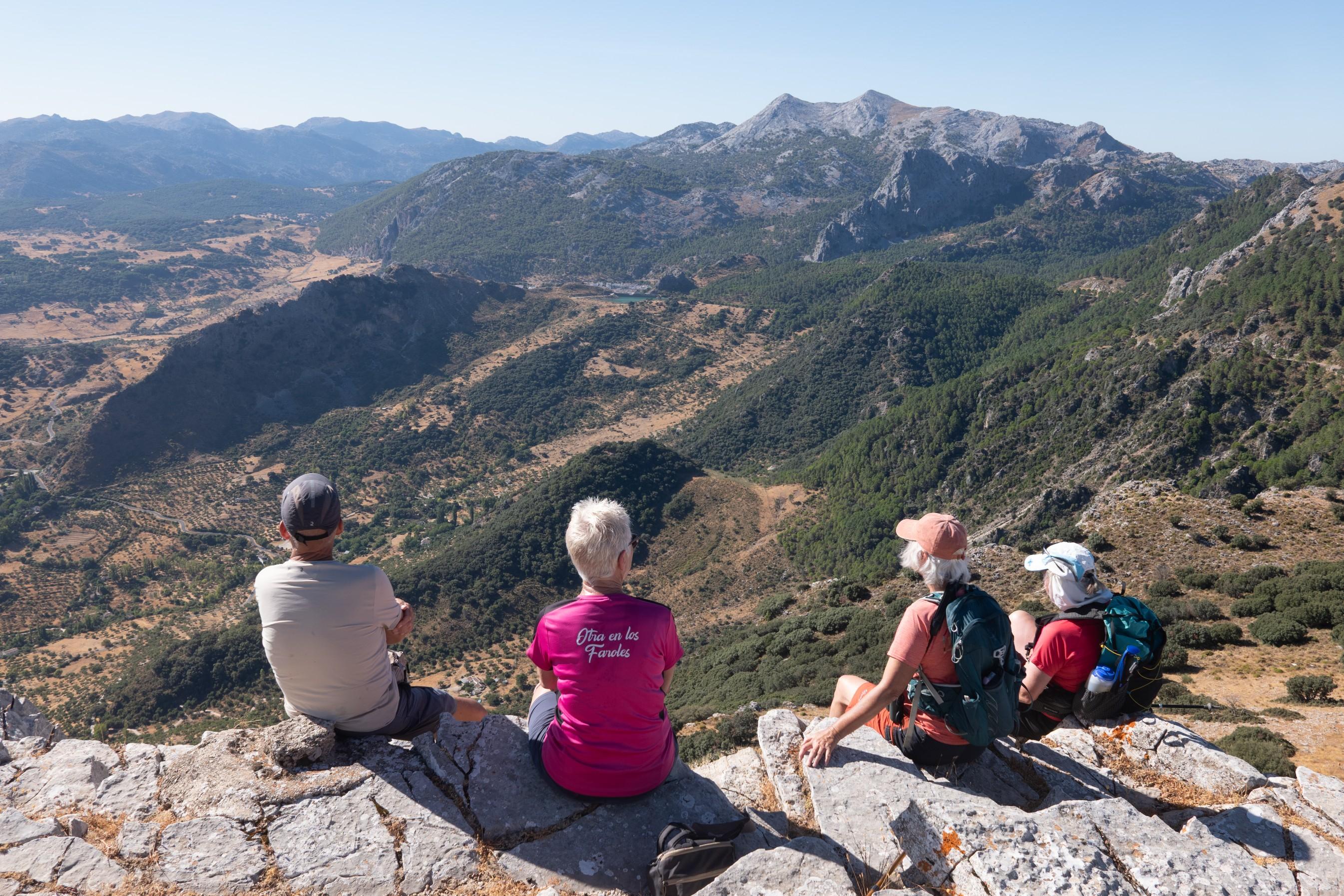 4 people in colourful tops sit at the summit of a mountain looking at a view to distant mountains, the Sierra de Grazalama