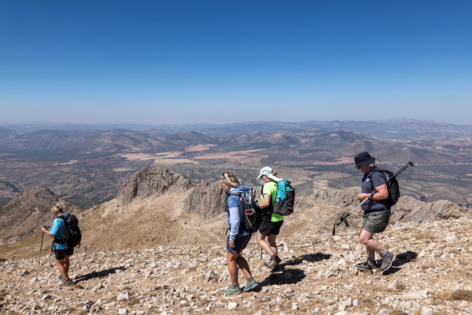 4 hikers walk along a high ridge with distant views beyond. Blue skies