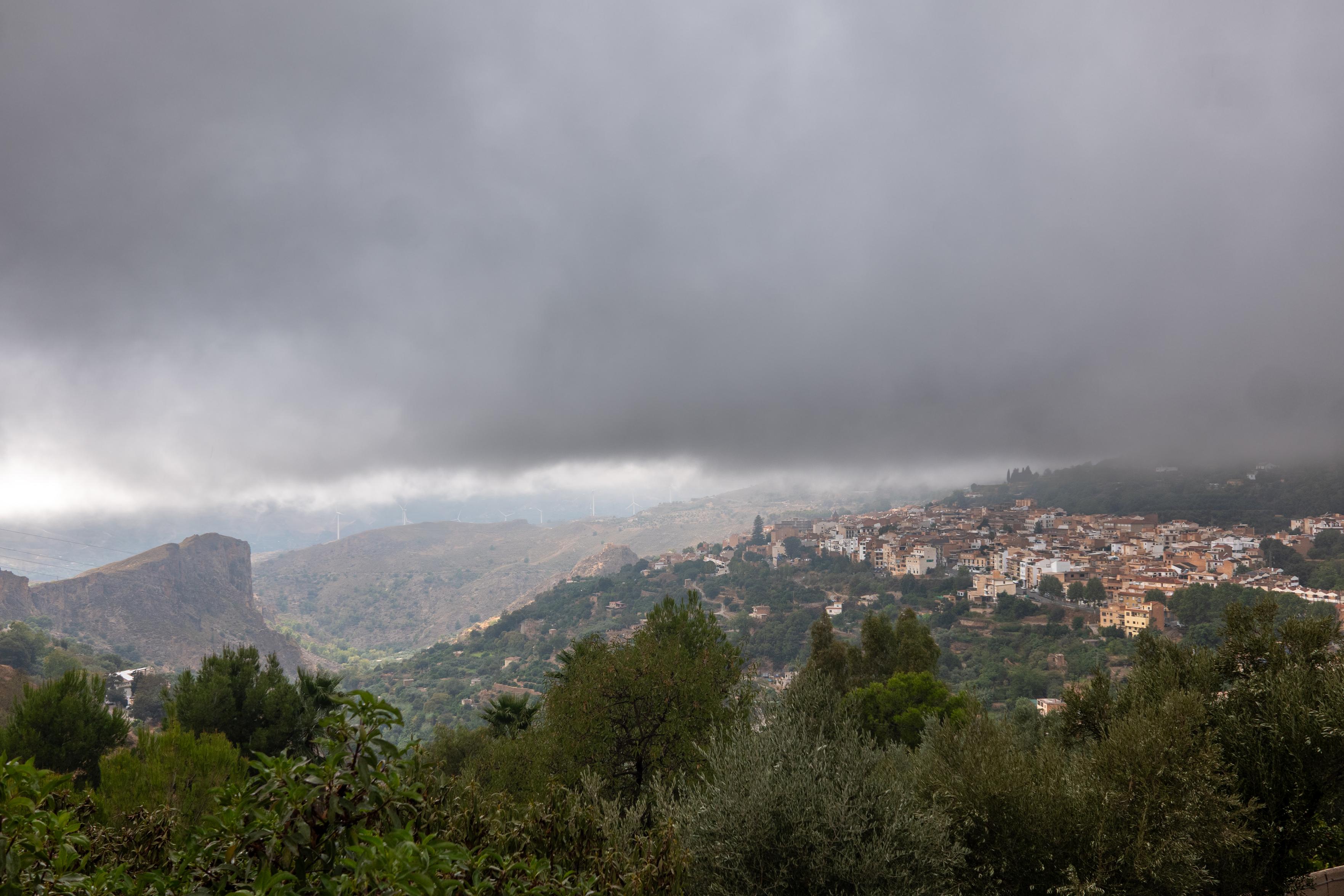 Below a grey dense cloud level a cliff to the left and a village to the right appears. Trees in the foreground