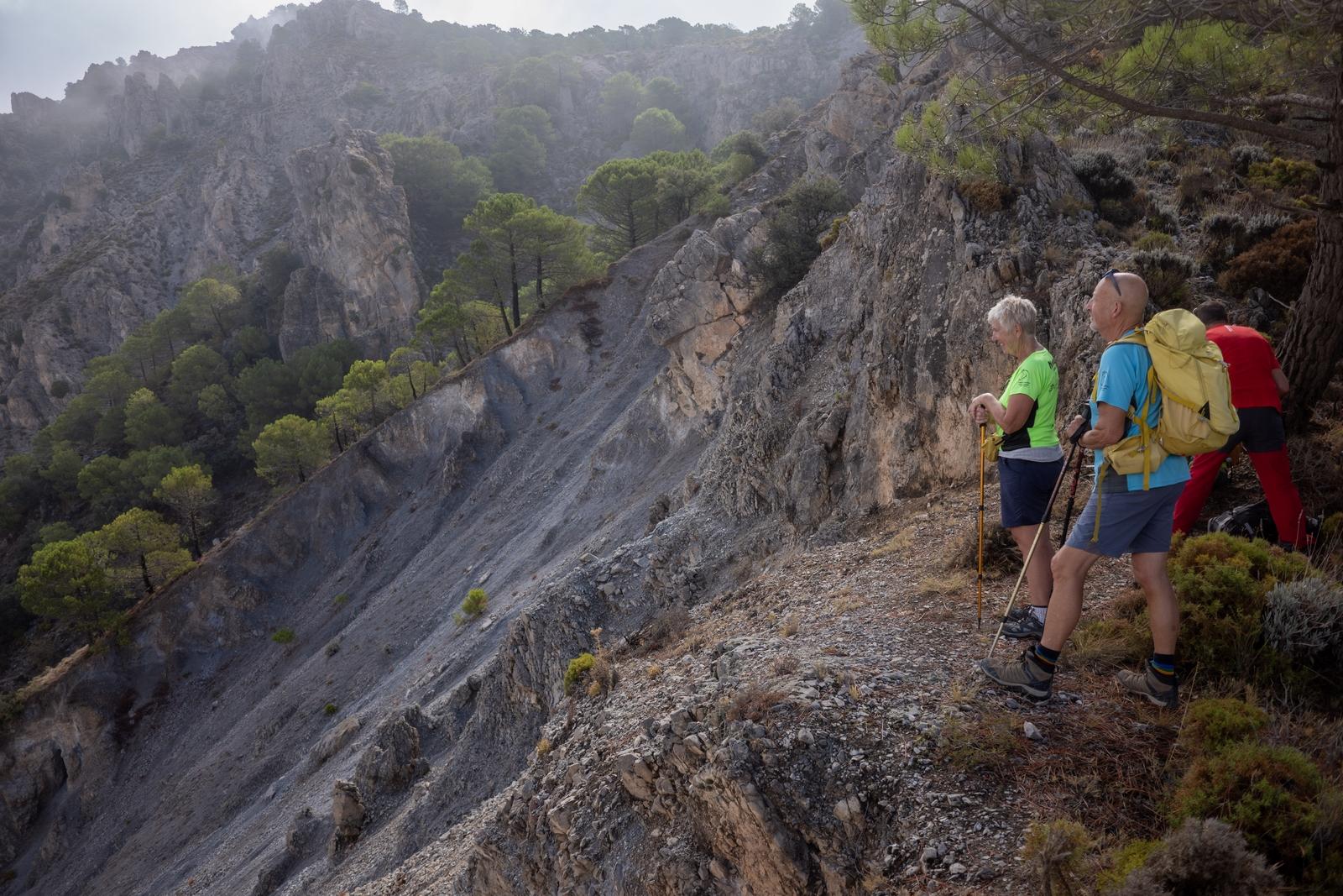 3 people on the right stand at a high viewpoint over an arid hillside with a few trees. Cloudier conditions higher