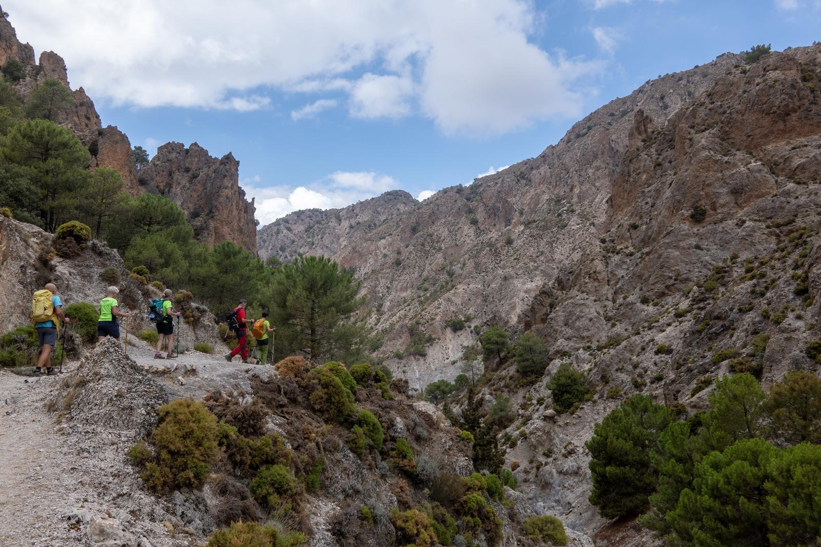 Hikers on a high mountain track on the right with a valley to their right and mountain beyond