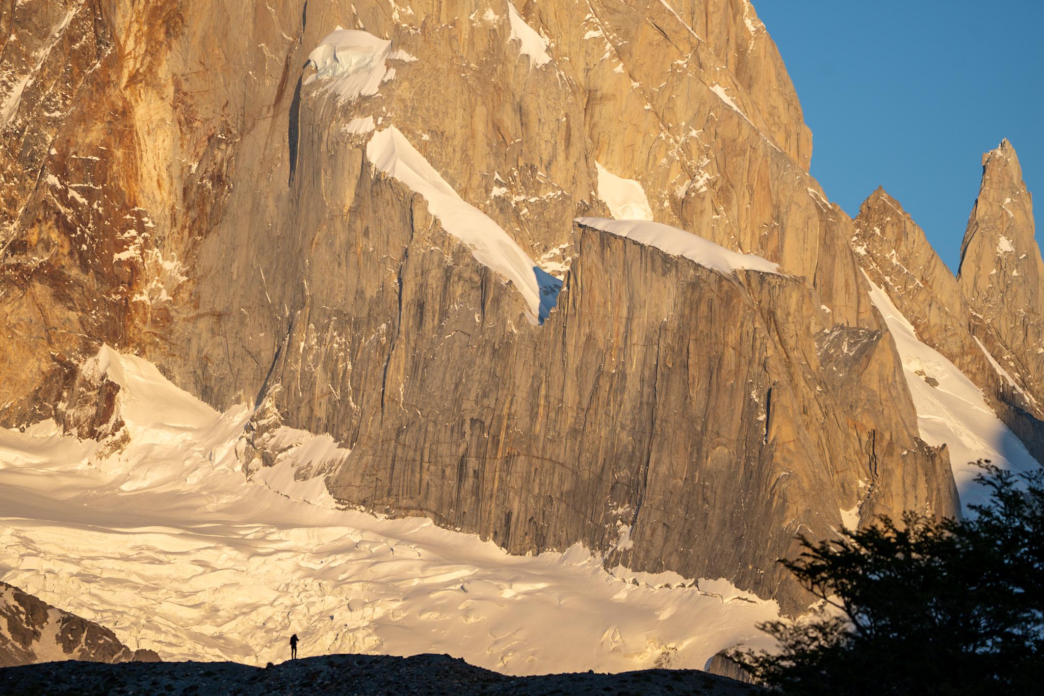 A person is silhouetted in the foreground whilst behind rises a sunlit wall of granite with glaciers and snowfields