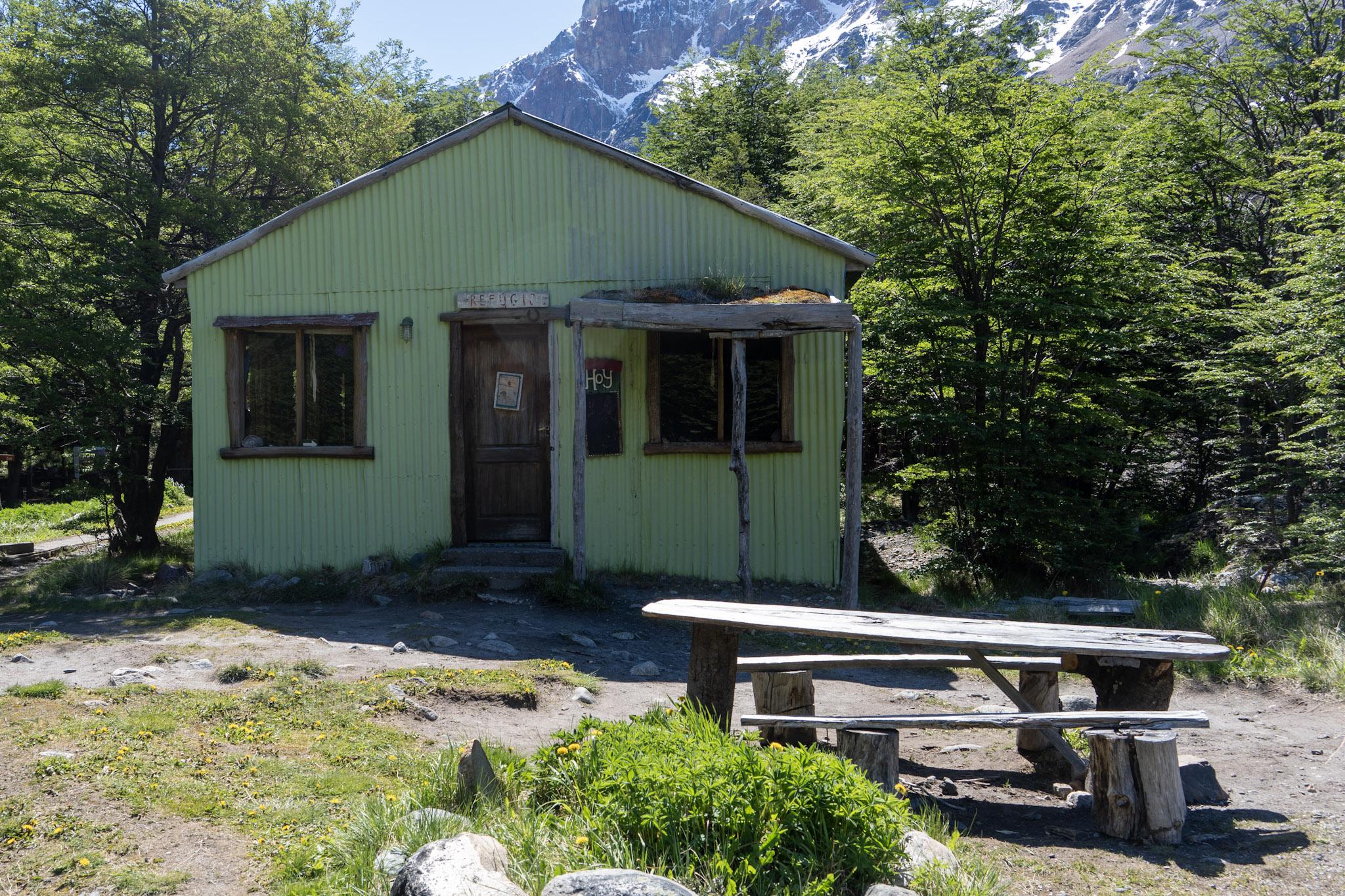 A green old building sits in a sheltered area of forest. It has a wooden table and benches outside