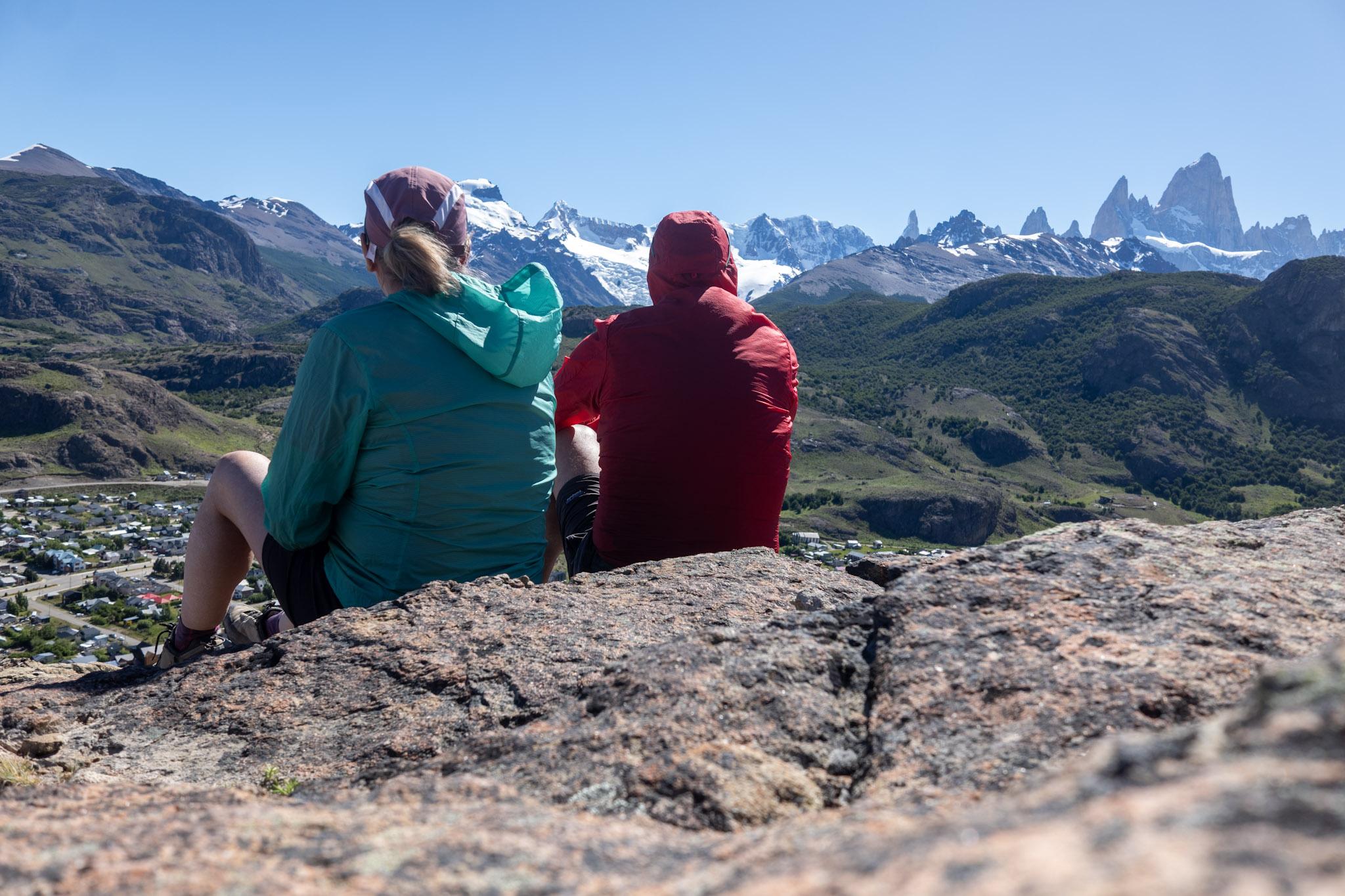 Two people sat looking out to some distance mountains. One in red, one in green jackets